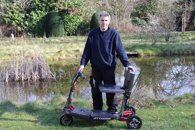 Man standing next to a mobility scooter by a pond.